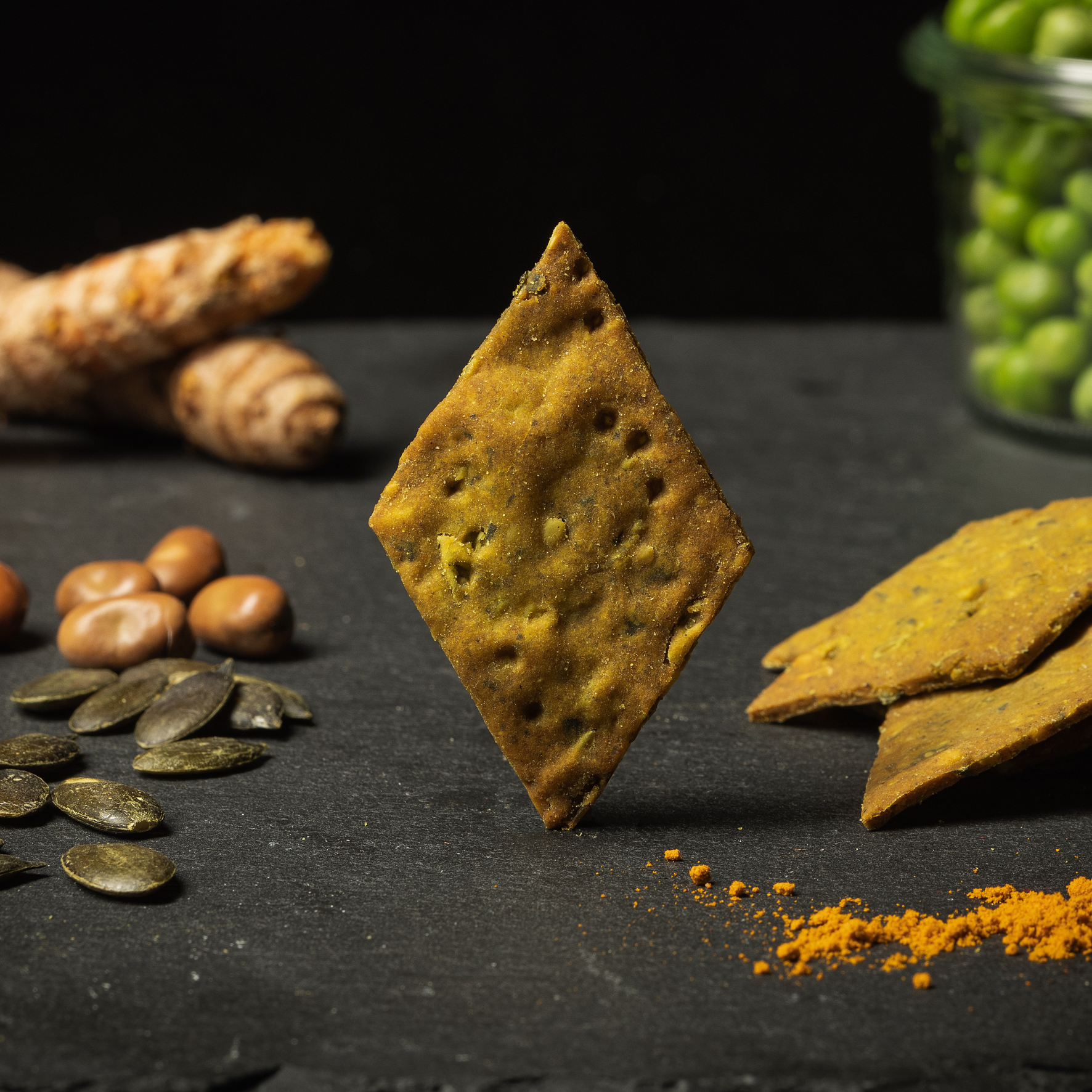 QiO Bio-Bohnencracker No.2 Kürbis-Kurkuma: Ein rautenförmiger, gebackener Snack, umgeben von Kürbiskernen und Kurkumawurzeln.